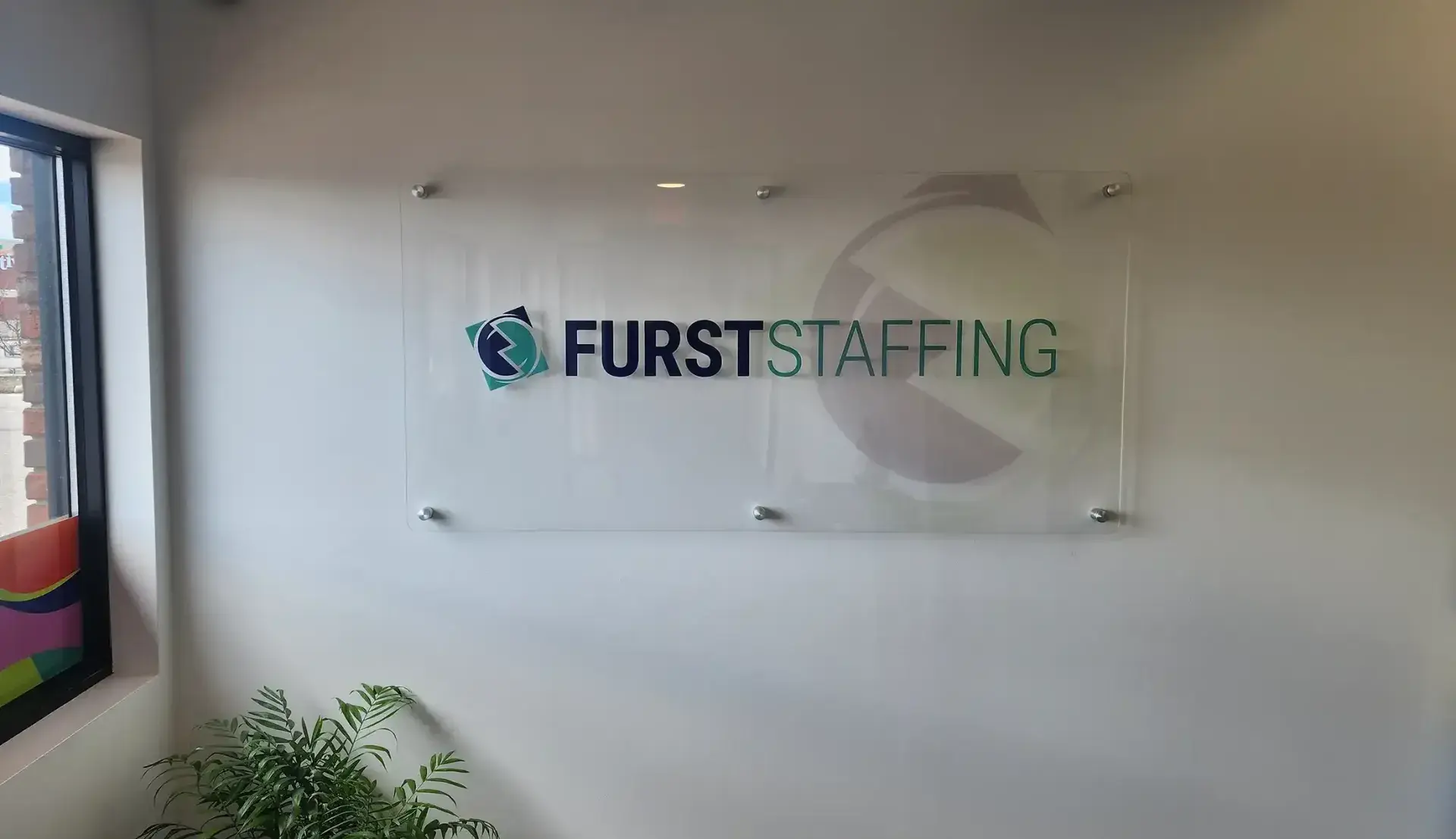 Office wall sign reading “FIRST STAFFING” on a white background, with a plant below and a framed print to the left