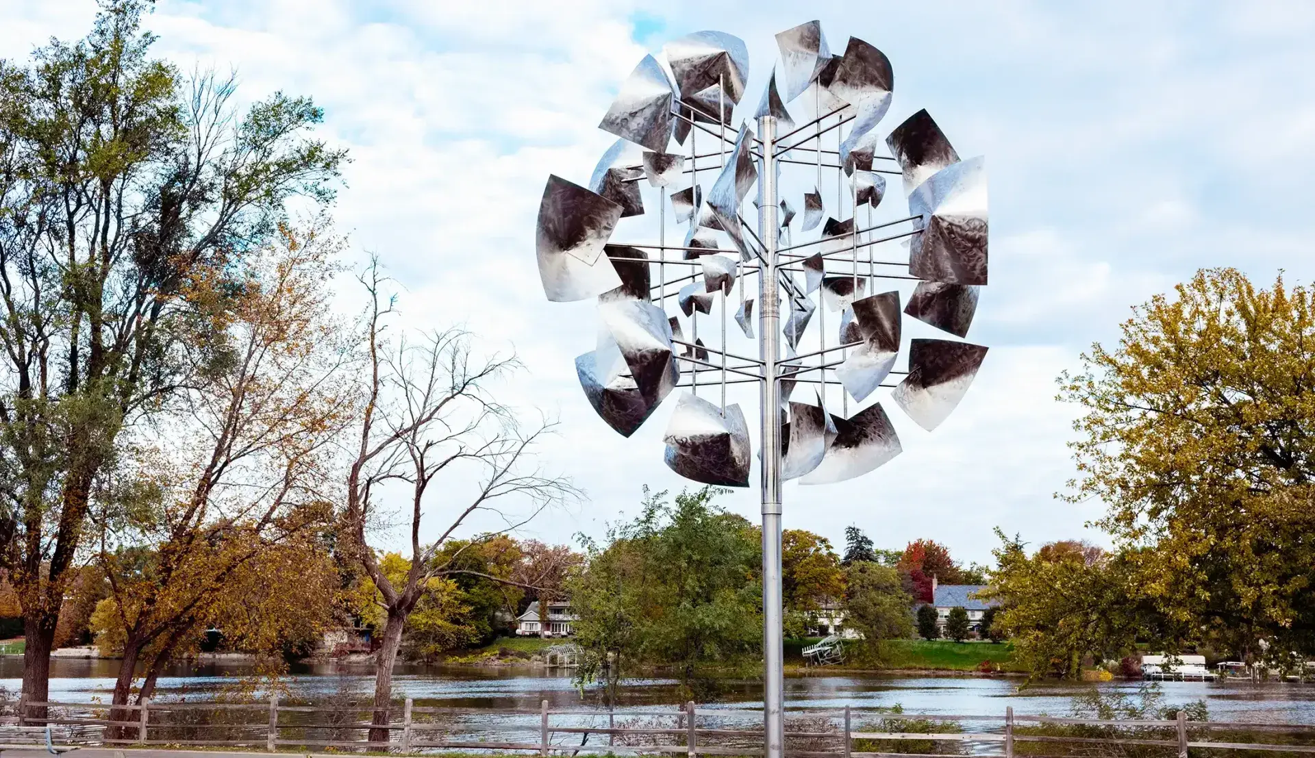 Outdoor kinetic wind sculpture with silver vanes beside a pond and autumn trees under a clear sky