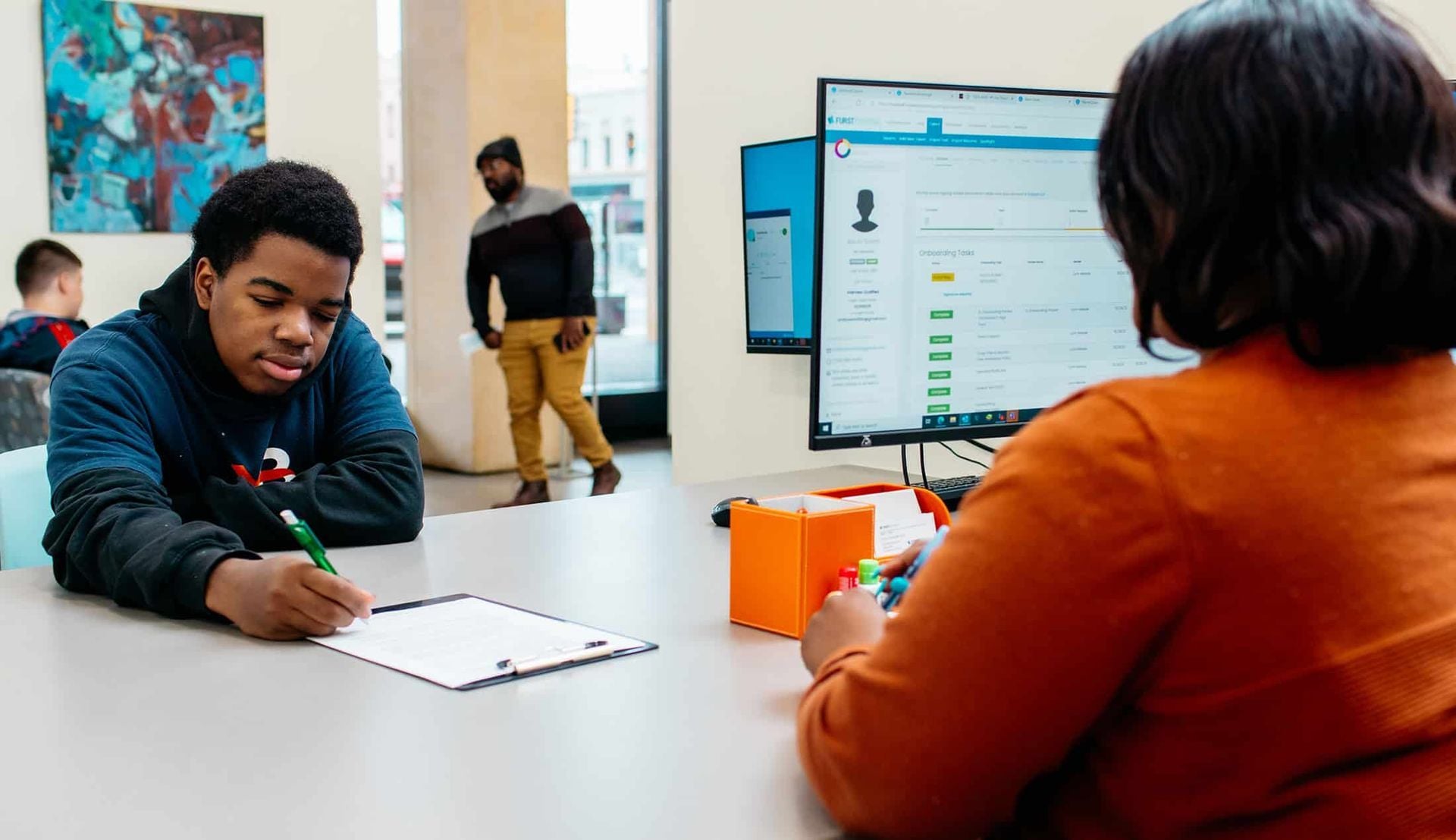 Two people talking across a table in a bright office, with a computer monitor between them.