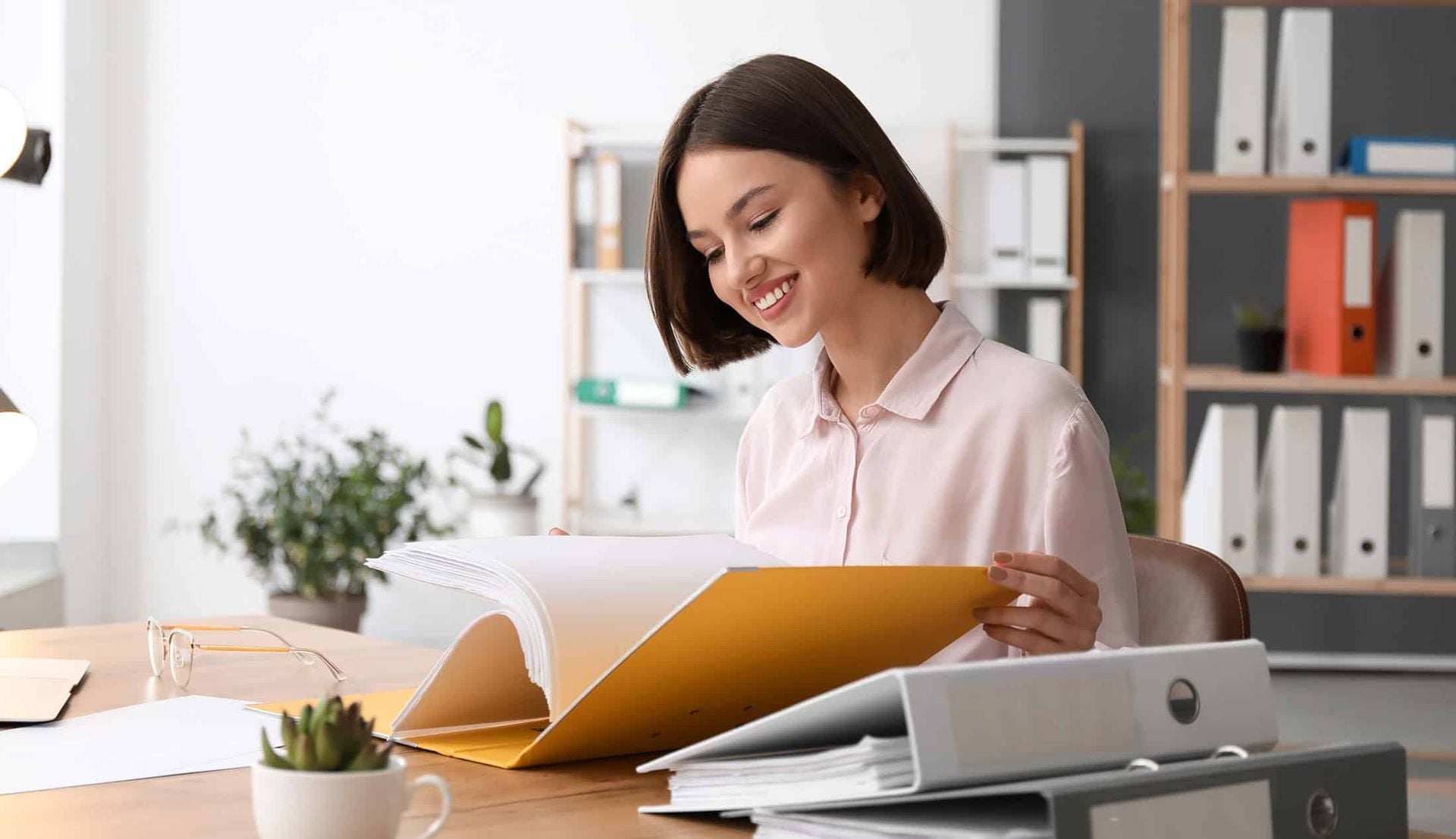 Woman smiling while reading papers at a desk in a bright office, with folders and a laptop nearby