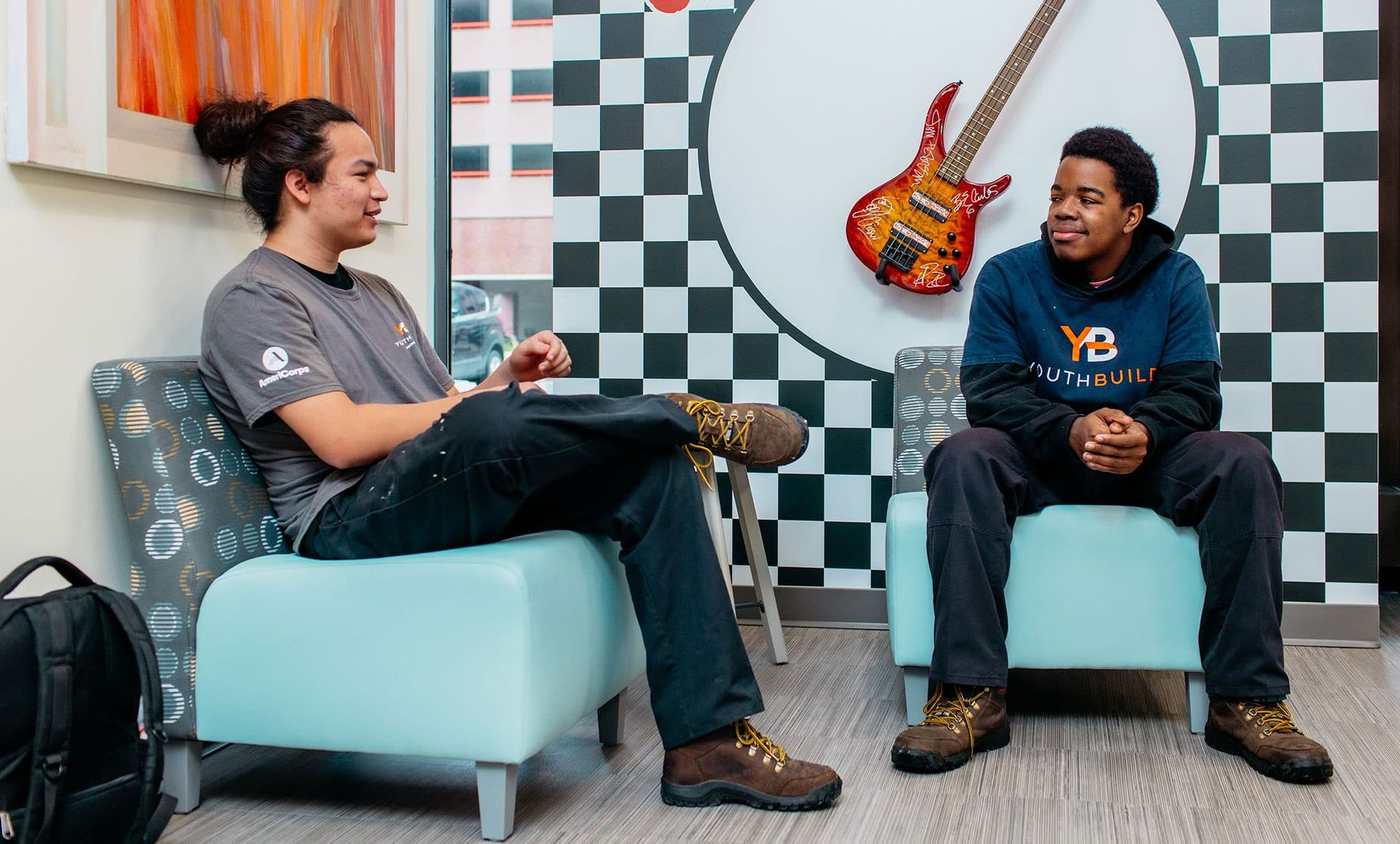 Two people seated in chairs talk in a checkered room with a colorful guitar on the wall.