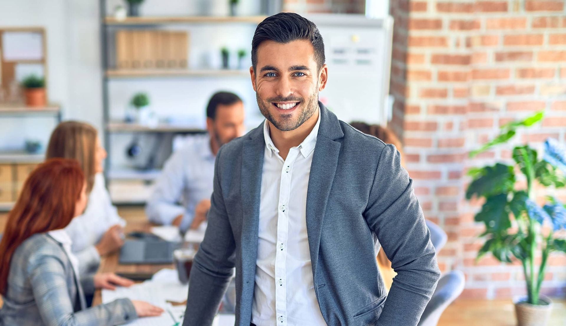 Smiling man in gray blazer in a bright office with coworkers in the background