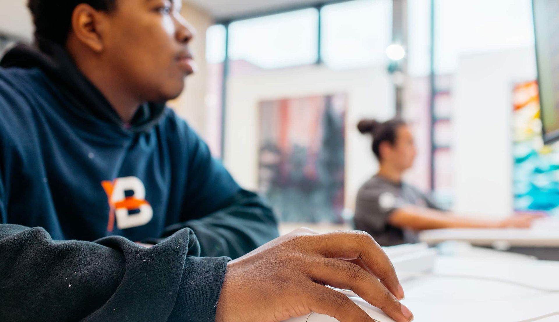 Student in a navy hoodie typing at a computer in a bright classroom, with others blurred in the background