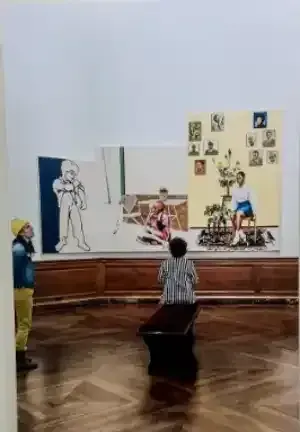 Person crouches in a museum gallery facing a wall-mounted art display with sketches and collage pieces.