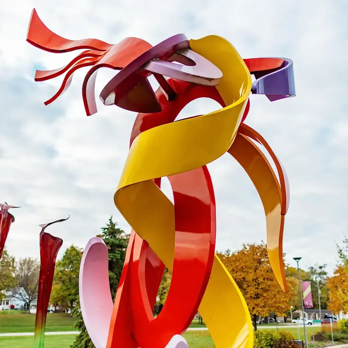 Abstract red, yellow, and purple ribbon-like sculpture in a park against a cloudy sky.