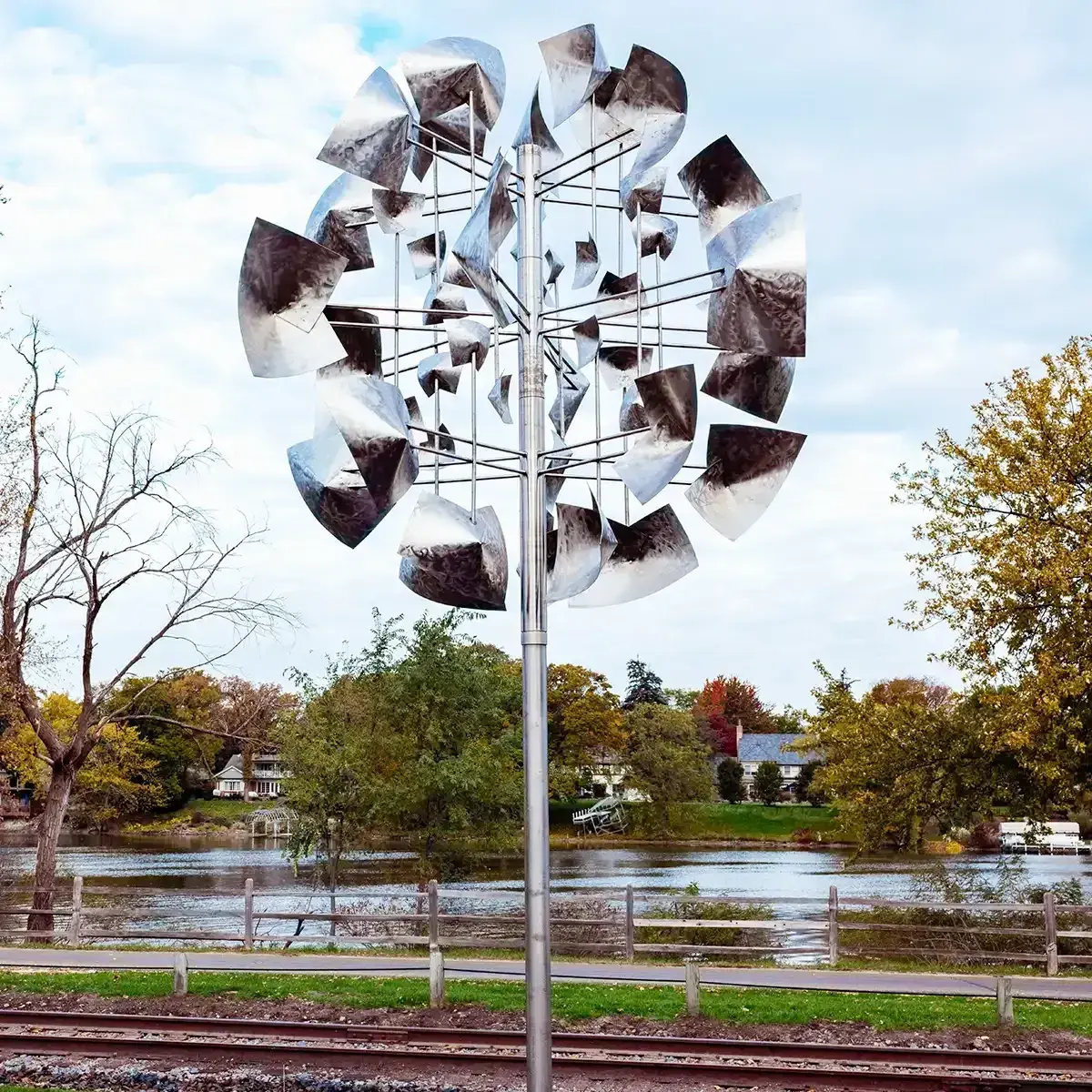 Metal wind sculpture by a riverside path with autumn trees and a cloudy sky